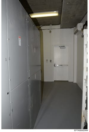 This photograph depicts an institutional or commercial interior hallway, featuring white and gray walls, fluorescent lighting, and multiple doors with safety signage. The space appears to be part of a building facility under investigation, with visib