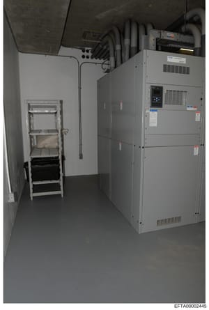 This photograph documents a utility/mechanical room within a property, showing industrial heating, ventilation, and air conditioning (HVAC) equipment along with electrical infrastructure. The image appears to be evidence documentation of building sys