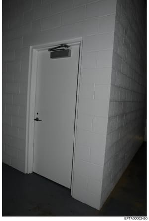This is a black and white photograph of a white-painted interior door with an automatic closer mounted above the frame. The door appears to be in a basement or institutional setting with concrete block walls. The photo was taken as part of evidence d