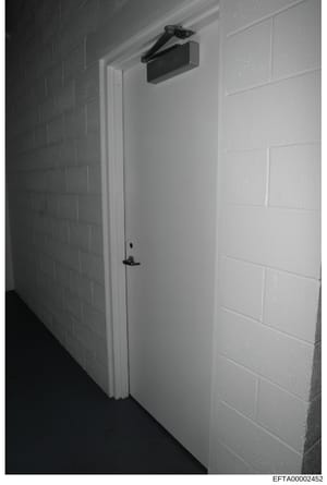 This is a black and white photograph showing an interior doorway with white painted walls and brick detailing. The door features a white frame, black handle hardware, and a black automatic door closer mounted at the top. The image appears to be evide