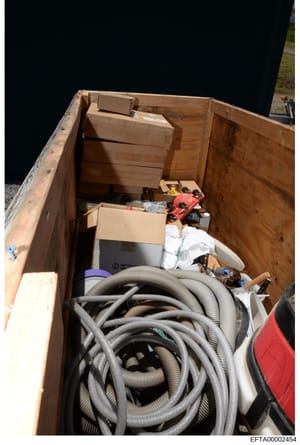 This is a photograph of the interior of a wooden storage container or dumpster filled with various household items, tools, and equipment. The contents include coiled hoses, cardboard boxes, power tools (visible red items), white linens or fabrics, an