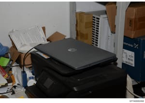 This photograph shows a desk or storage area containing a black HP laptop, stacked cardboard boxes, papers and documents, and various office supplies. The image appears to be evidence documentation from a search or seizure, showing the physical envir
