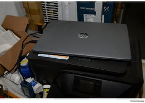 This is a photograph of computer equipment seized as evidence, showing an HP laptop positioned on top of a black printer, with various cables and supplies visible in the background. The image documents the physical state of electronic devices during 