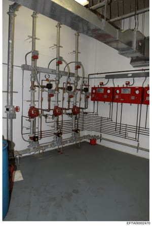 Photograph of an installed fire suppression and sprinkler system in what appears to be a basement or utility room. The image shows multiple vertical copper piping columns with red manual shut-off valves, red electronic control boxes mounted on the wa