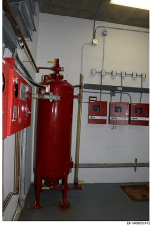 Photograph of a building's fire suppression system room showing a large red pressurized tank, fire alarm control panels, and associated piping and electrical infrastructure. This image appears to be evidence documentation from a property inspection, 
