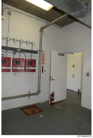 This photograph shows a basement or utility room interior featuring fire alarm panels, emergency fire extinguisher, exposed piping, and utility infrastructure. The image appears to be from a property inspection or evidence documentation related to th