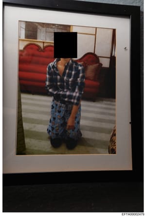 This is a framed photograph showing the interior of a residence with distinctive red upholstered furniture. A person is visible wearing a blue and white plaid/checkered shirt and patterned skirt, with their face redacted by a black square. The settin