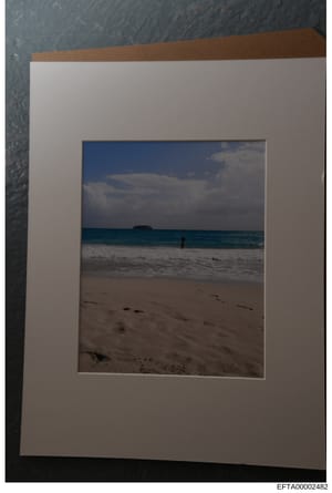 This is a color photograph of a tropical or subtropical beach scene, mounted on white matting with a brown backing. The image shows a sandy beach with calm turquoise waters, white clouds in a blue sky, and what appears to be a small island or landmas