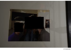 This is a photograph of an interior room showing multiple individuals whose faces have been redacted with black rectangles. The setting appears to be a residential space with beige/tan walls, doorways, and decorative elements including framed artwork