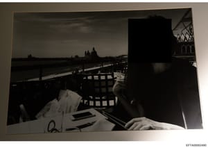 This is a black and white photograph showing a person seated at a dining table on what appears to be a waterfront terrace or balcony overlooking Venice, Italy. The image captures a scenic vista with the Grand Canal, iconic Venetian architecture inclu