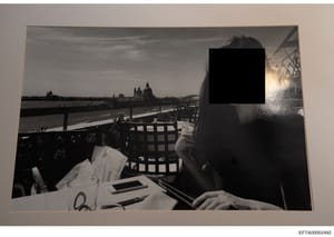 Black and white photograph showing a woman seated at an outdoor waterside dining venue. The image captures a scenic European location with distinctive architecture including domed buildings visible across the water, consistent with Venice, Italy. The
