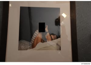 This is a mirror selfie photograph showing a person in a bedroom setting taking their own picture. The subject's face has been redacted with a black rectangle. The image appears to be documentary evidence collected during the investigation, preserved