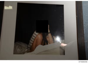 This is an evidence photograph from the Epstein investigation files showing a person with their face obscured by a black square. The image appears to depict an individual on a bed or similar surface wearing striped clothing. The obscuring of the face