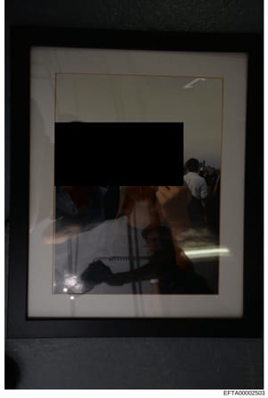 This is an evidence photograph showing the interior of a room with multiple individuals present. The image appears to be taken through or reflected in glass, showing at least two people in what appears to be a residential or institutional interior sp