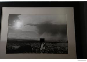 This is a black and white photograph showing a figure standing in an arid, desert landscape with sparse vegetation and flat terrain extending to the horizon. The person's head is redacted with a black box. The photo appears to be evidence documentati