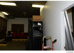 This is an evidence photograph showing an interior storage or equipment room containing a tall black server/equipment rack, red storage containers, a cardboard box, and a red hand truck. The room has fluorescent ceiling lights, white and dark walls, 