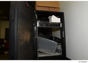 This is an evidence photograph showing the interior of a black server rack or equipment cabinet containing various networking and computing hardware, including server units with indicator lights, cables, and storage containers. The photo appears to b