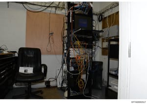 This photograph shows an interior space containing networked computer equipment, servers, and extensive cabling. The room appears to be a makeshift server or equipment storage area with multiple devices stacked vertically, numerous colored network ca