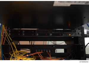 This photograph shows a server rack or network equipment installation containing multiple rack-mounted devices with status indicator lights, network connections, and extensive cable management with yellow and orange network/power cables. The image ap