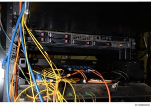 This is a photograph of a server rack containing multiple hard drive bays, network equipment, and extensive cable management. The image shows professional IT infrastructure with color-coded cables (yellow, blue, orange) and numbered rack positions, l