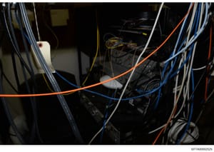 This is a photograph of the interior of technical equipment or a server/networking cabinet, showing multiple colored cables (orange, blue, white, yellow) connected to various black electronic devices and components. The image appears to be documentat