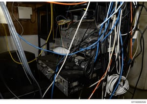 This is a photograph of a server room or network equipment closet showing multiple computer servers, networking hardware, and extensive cabling in various colors (blue, yellow, orange, white, black). The image documents the physical infrastructure an