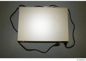 This is a photograph of a rectangular electronic device with a cream/beige colored top surface and black power cord attached. The device appears to be some form of lighting equipment or electronic apparatus, photographed against a gray wall. The imag