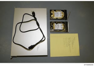 This evidence photo documents seized computer equipment including two hard disk drives, a power cable, and a handwritten yellow inventory note. The items appear to be evidence collected during the investigation, with the hard drives bearing manufactu