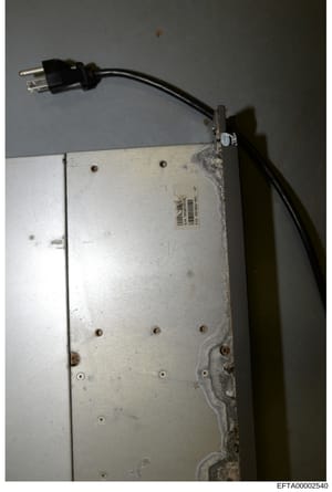 This is a forensic evidence photograph showing a white electronic device or equipment with a black power cord attached. The device appears weathered with visible dust and wear marks, and bears what appears to be an identification label or barcode on 