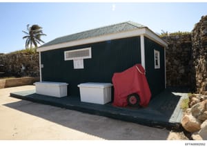 This is a photograph of a small residential building or outbuilding with dark green corrugated metal siding, white trim, and a pitched roof. The structure sits in an arid or semi-arid coastal location (indicated by palm trees and stone walls). In fro