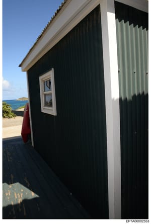 This photograph shows an exterior view of a small black and white structure with modern architectural features, located on a beachfront property with clear ocean views. The image captures a wooden deck, white-trimmed windows, and corrugated metal sid