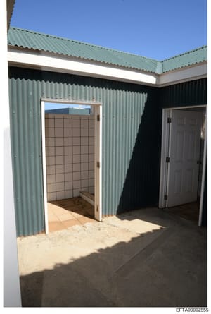 This photograph depicts the interior of a small corrugated metal structure, likely a residential outbuilding or accessory structure. The image shows a tiled bathroom or utility room with an open doorway on the left and a closed door on the right. The