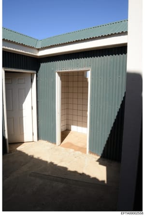 This is a photograph of a simple metal building with corrugated walls and roof. The structure shows two doorways—one white six-panel door on the left and one open doorway on the right leading to an interior room with tiled walls. The building appears