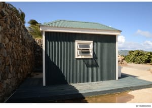 This photograph shows a small, modern residential structure with corrugated metal siding and a green metal roof located on what appears to be a coastal property. The building features a wooden deck entrance and a single window. The surrounding landsc