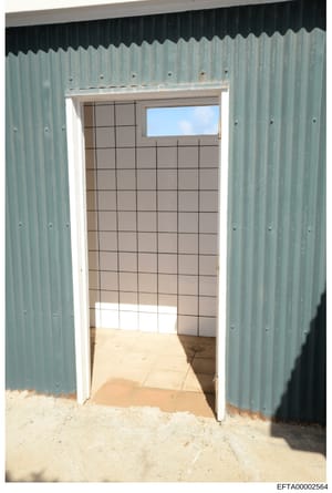 This is a photograph showing the interior of a corrugated metal building or structure, taken through an open doorway. The image reveals a room with beige/tan tiled walls in a grid pattern, a concrete floor with sand/dirt visible at the entrance, and 