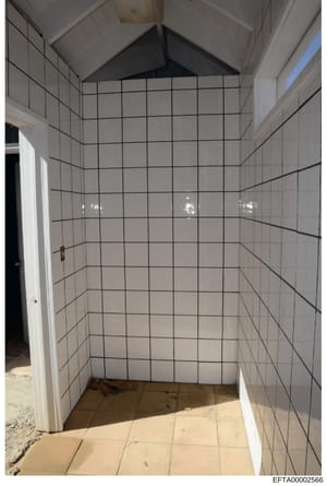 This photograph depicts an interior bathroom room with white tile walls and a vaulted ceiling. The room appears to be under construction or in a state of renovation, with minimal fixtures visible and exposed construction materials on the floor. The B