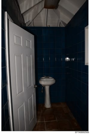 This is a photograph of a bathroom interior, showing a pedestal sink against walls covered in dark blue tile. The room features a white six-panel door, exposed ceiling with ductwork, and brown flooring. The image appears to be documentation of proper