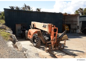 This photograph shows a property or facility containing heavy construction and industrial equipment, including a JLG telehandler, a CAT skid loader, and an orange loader vehicle, situated in front of dark metal storage buildings with corrugated metal