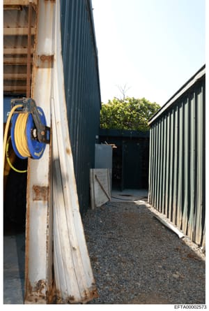 This photograph shows the exterior of an industrial or warehouse building featuring corrugated metal structures, a blue and yellow hydraulic hose reel mounted on a weathered white panel, gravel flooring, and metal fencing. The image appears to be doc