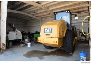 This photograph depicts the interior of an industrial storage or maintenance facility containing a yellow Caterpillar (CAT) compactor machine, along with various other equipment and tools. The image shows a corrugated metal structure with exposed bea