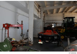 This is a photograph of an industrial storage facility or garage interior containing various pieces of heavy equipment and machinery. The space shows a red hydraulic press on the left, a red and black jet ski or personal watercraft in the center, a C