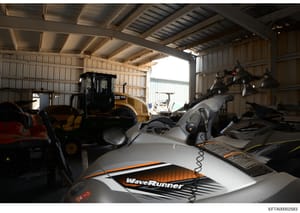 This photograph shows the interior of a large metal storage warehouse or hangar containing multiple personal watercraft (jet skis), including a prominent WaveRunner model in the foreground, along with heavy construction equipment including a CAT load
