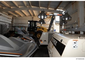 This photograph documents the interior of a large metal warehouse or storage facility containing various equipment and vehicles. The space shows a CAT (Caterpillar) wheel loader, a WaveRunner jet ski, industrial lighting fixtures, and other storage e