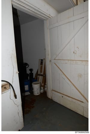 This photograph depicts the interior of a storage or utility room, showing white paneled walls, various equipment including what appears to be a blue container and white bucket, and evidence of wear and staining. The image was taken as part of the in