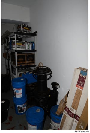 This photograph shows a residential or commercial storage area containing multiple blue drums (likely motor oil or similar automotive fluids), black storage containers, and various maintenance supplies arranged on metal shelving. The image appears to