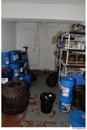 This is a photograph of a storage or utility room containing multiple large industrial drums (blue and black), automotive fluids, oils, shelving units with product boxes, a tire, and various maintenance supplies. The room shows signs of age with stai