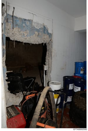 This is an evidence photograph showing an interior storage or basement space with various equipment, tools, and supplies. The image depicts a deteriorating wall with exposed drywall, a large opening or doorway leading to an adjacent dark room, and nu