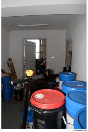 This is a photograph of an interior storage or utility room containing multiple industrial storage drums and containers in blue and red colors, along with various tools and equipment. The room appears to be a basement or utility space with white wall