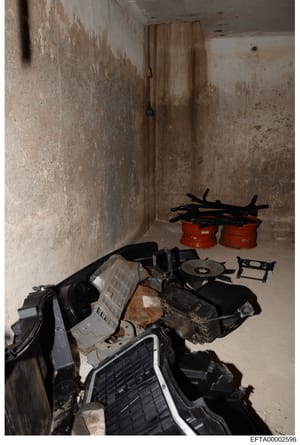 This is an evidence photograph from the Epstein investigation showing the interior of a concrete storage room or basement space containing various seized items including automotive parts, electrical equipment, red containers, and black materials. The