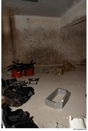 This is an evidence photograph showing an interior room or basement space containing various items including red containers, cardboard boxes, black equipment/machinery parts, and metal fixtures. The room shows signs of significant moisture damage and